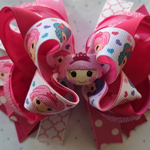 May include: A large, layered hair bow in shades of pink. The bow features a central doll figure with a pink crown. The ribbon includes a repeating pattern of doll faces and hearts. The bow is approximately 6 inches wide.