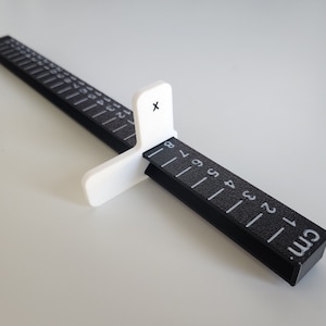 Peut inclure: Une règle en plastique noire avec un marqueur coulissant blanc. La règle mesure en centimètres et comporte des graduations jusqu'à 10 cm. Le marqueur porte un 'X' imprimé.