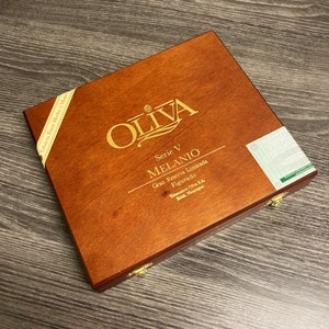 Puede incluir: Una caja de madera con una etiqueta dorada que dice "OLIVA Serie V MELANIO Gran Reserva Limitada Figurado Tabaccos Oliva S.A. Estelí, Nicaragua".