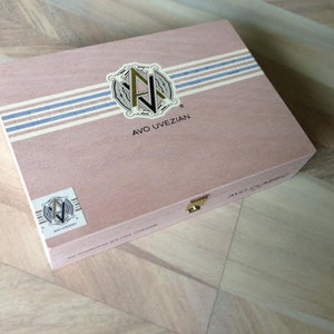 May include: A wooden box with a pink lid and blue and white stripes. The box has a gold latch and a gold logo with the text "AVO UVEZIAN".