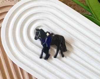 May include: Black enamel pin featuring a person in a blue jacket leading a black horse. The pin is on a white background with a textured surface.