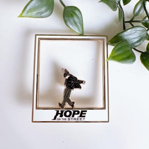 Peut inclure: Épinglette en émail dorée représentant une silhouette noire d'une personne dansant avec le texte "HOPE ON THE STREET" en dessous. L'épingle est dans un cadre blanc avec une bordure dorée.