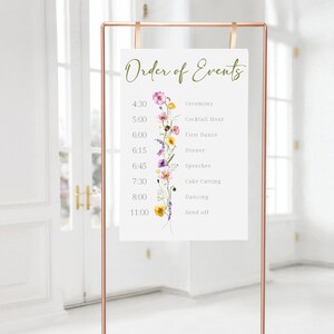 May include: A white sign with a floral design and the text "Order of Events" with a list of wedding events and times. The sign is on a gold stand.