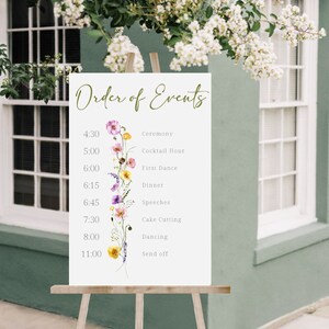 May include: A white sign with the words "Order of Events" written in green script. The sign lists the order of events for a wedding reception with times and descriptions, such as "Ceremony", "Cocktail Hour", "First Dance", "Dinner", "Speeches", "Cake Cutting", "Dancing", and "Send off". A floral illustration with pink, purple, yellow, and white flowers decorates the left side of the sign.