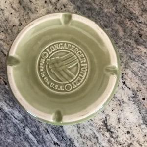 Puede incluir: Un cenicero redondo verde claro con borde blanco. El centro presenta el texto "Longaberger Pottery" y "Made in the U.S.A." en un diseño circular. Tiene cuatro ranuras pequeñas para cigarrillos.