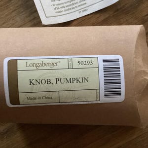 May include: A brown paper package with a white label featuring the text Longaberger, 50293, and KNOB, PUMPKIN. A separate instruction sheet includes the text TO REMOVE (OR REPLACE) KNOB, Do not twist knob on or off of lid. This may cause marring of lid surface.