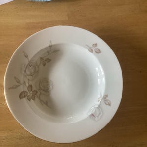 Puede incluir: Un cuenco de cerámica blanca con un delicado patrón floral de rosas y hojas de color gris claro en el borde. El cuenco tiene un diseño circular liso con un sutil borde dorado en el exterior, lo que le confiere un aspecto limpio y sencillo.