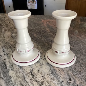 May include: Two white ceramic candle holders with a flared base and a wide top. Each holder has a decorative burgundy trim and floral accents. The candle holders are displayed on a speckled countertop.
