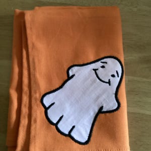 May include: A folded orange kitchen towel featuring a white ghost appliqué with a black embroidered outline. The ghost has a simple smiling face with two eyes and a curved mouth, positioned on the lower right side of the fabric surface.