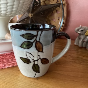 May include: A ceramic coffee mug with a brown rim and handle. The mug has a light blue, white, and brown glaze with a leafy design.