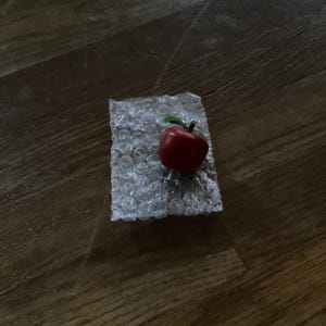 May include: A small, red, apple-shaped object with a brown stem and a single green leaf, resting on a rectangular piece of clear, textured bubble wrap packaging material.