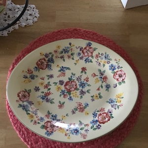 May include: An oval, cream-colored serving platter with a floral pattern of red, blue, and yellow flowers. The platter sits on a round, red braided placemat. The design is a classic, vintage style.