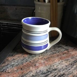 May include: White ceramic mug with a blue interior and horizontal blue and gray stripes. The mug has a curved handle and is set on a granite countertop. Kitchenware.