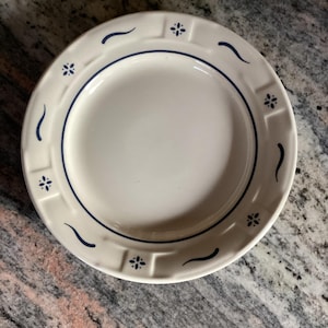 May include: Off-white ceramic plate with a blue floral and wavy design. The plate has a blue border and a decorative rim with small blue flower and wave motifs. The plate is round and appears to be new.