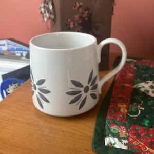 May include: White ceramic mug with a black floral design. The mug has a handle and is sitting on a wooden surface.