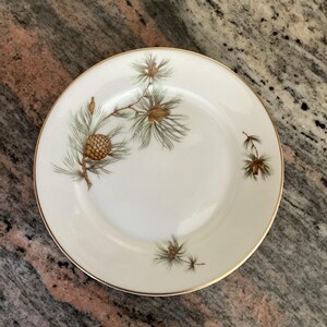 May include: A white ceramic plate with a gold rim, featuring a detailed illustration of a pine branch with a pinecone and needles in shades of brown and green. The plate is round and has a classic design.