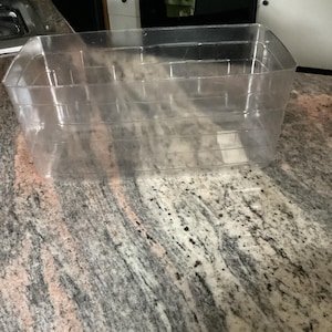 May include: A transparent plastic storage container, rectangular in shape, with internal dividers. It is empty and placed on a granite work surface. Ideal for organizing items, the container is clear.