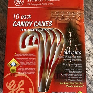 Puede incluir: Una caja de 10 luces de bastón de caramelo GE Holiday Classics. La caja es roja con letras blancas y una imagen de las luces de bastón de caramelo. Las luces tienen 5,5 metros de largo y 50 luces. La caja dice "White Cane/Red Stripes Indoor/Outdoor".