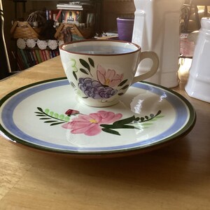 Könnte beinhalten: Ein handbemaltes Tee-Set. Die Tasse und Untertasse sind weiß mit einem Blumendesign in Rosa, Lila und Grün. Die Untertasse hat einen blauen und grünen Rand. Das Set steht auf einem Holztisch.