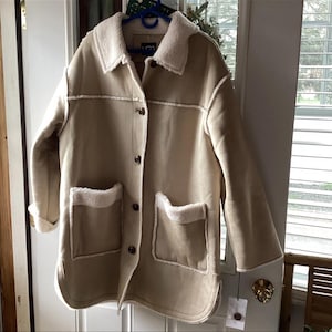 May include: A tan-colored coat with a cream-colored shearling collar and trim. The coat has two large front pockets and is buttoned up. The UGG logo is visible on the hanger. The coat is hanging on a door.