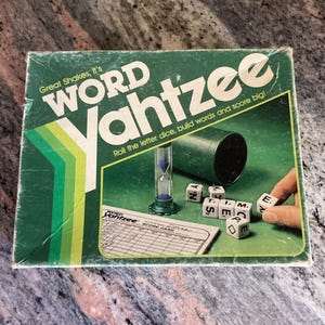 Peut inclure: Une boîte de jeu Word Yahtzee vintage avec du texte vert et blanc. Le jeu comprend des dés de lettres, un sablier et une carte de score. La boîte indique "Great Shakes, it's Word Yahtzee. Roll the letter dice, build words and score big!"