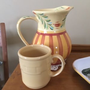 Puede incluir: Un juego de jarra y taza de cerámica. La jarra tiene una parte superior de color crema con diseños de hojas verdes y azules y un cuerpo a rayas rojas y amarillas. La taza es de color crema sólido con una textura similar a ladrillos.