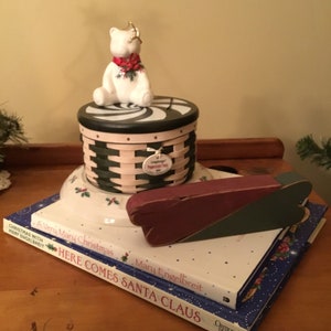May include: A white ceramic polar bear figurine with a red and green bow sits on top of a black and white striped lid of a green and white wicker basket. The basket has a tag that says "Longaberger Holiday Memories". The basket is sitting on a white plate with green holly leaves. There are two books stacked on top of the plate. The top book is titled "A Very Mary Christmas" by Mary Engelbreit. The bottom book is titled "Here Comes Santa Claus" by Mary Engelbreit.