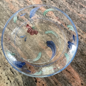 May include: Clear glass plate with a blue rim and painted floral and leaf designs in shades of blue, green, and red. The plate has a circular shape and is suitable for serving food.