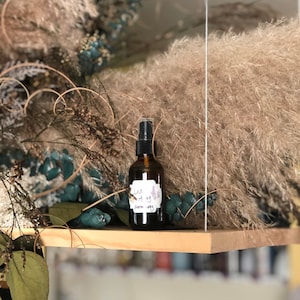 May include: A brown glass bottle with a black spray nozzle and a white label that says "Luck & Joy" and "Garden Spray". The bottle is sitting on a wooden shelf with dried flowers and plants in the background.
