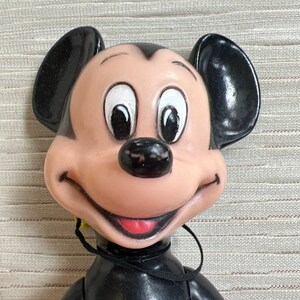 May include: A vintage Mickey Mouse doll with a smiling expression. The doll features a black body, large black ears, and a tan face with white eyes. The doll has a red shirt and a black string around its neck.