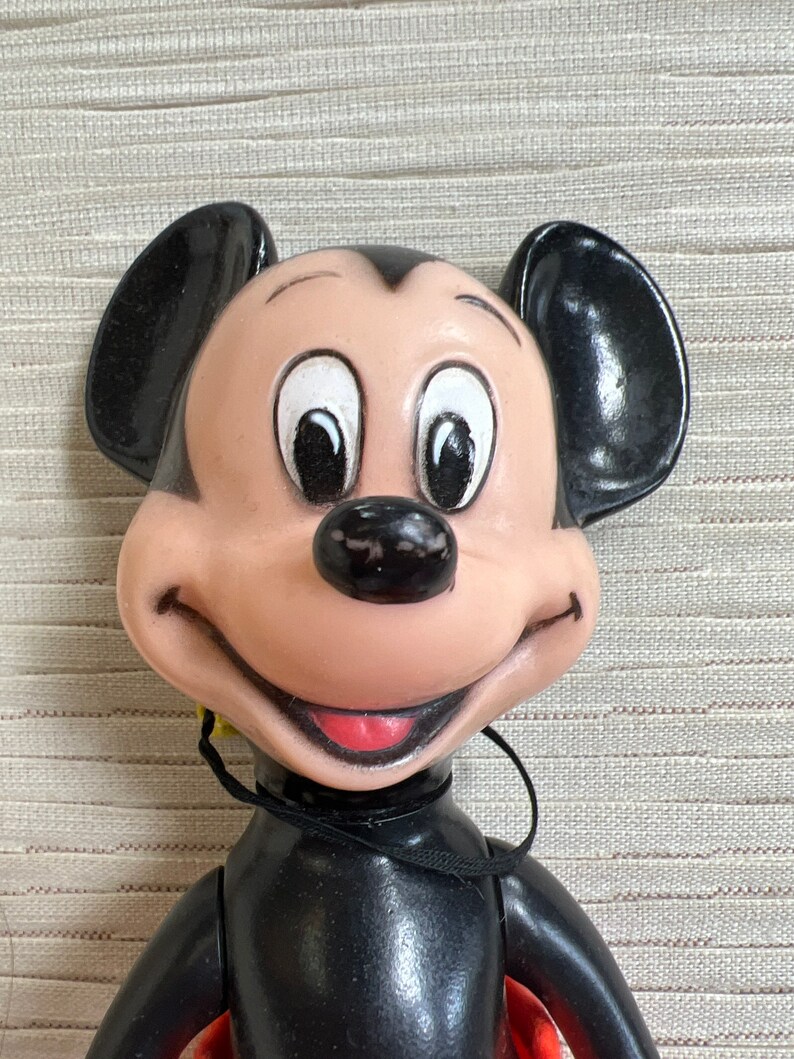 May include: A vintage Mickey Mouse doll with a smiling expression. The doll features a black body, large black ears, and a tan face with white eyes. The doll has a red shirt and a black string around its neck.