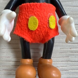 May include: A vintage Mickey Mouse figurine. The figure has a black head, arms, and legs, with white gloves and brown shoes. It wears a red felt shorts with two yellow circles.