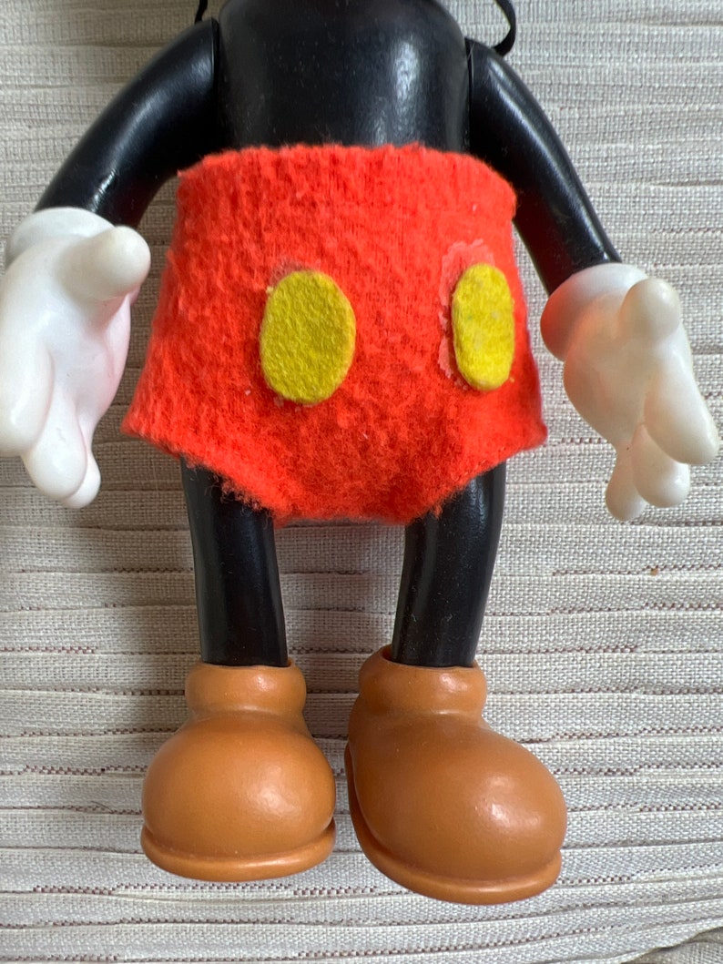 May include: A vintage Mickey Mouse figurine. The figure has a black head, arms, and legs, with white gloves and brown shoes. It wears a red felt shorts with two yellow circles.