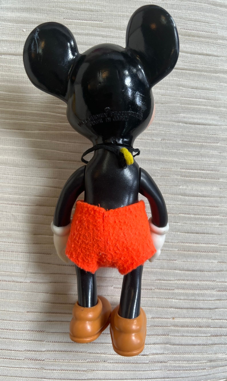 May include: Vintage Mickey Mouse figurine, viewed from the back, with black body and ears, orange shorts, and tan shoes. The figure has white gloves and a black collar with a yellow accent. The text "WALT DISNEY PRODUCTION MADE IN JAPAN" is visible.