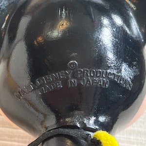 May include: Black Mickey Mouse figurine with visible text: "WALT DISNEY PRODUCTIONS MADE IN JAPAN". The figurine has a black body, pink face, and yellow accents. The image is a close-up of the back of the figurine.