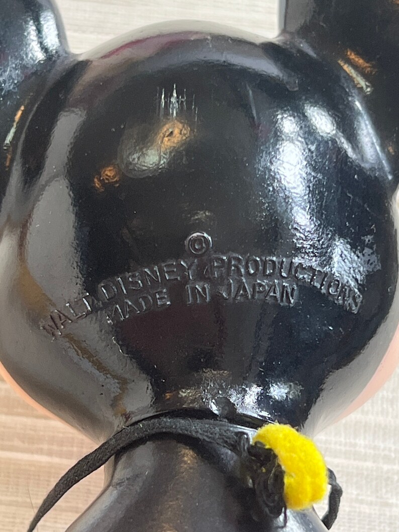 May include: Black Mickey Mouse figurine with visible text: "WALT DISNEY PRODUCTIONS MADE IN JAPAN". The figurine has a black body, pink face, and yellow accents. The image is a close-up of the back of the figurine.