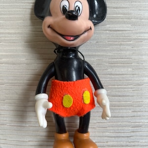May include: A vintage Mickey Mouse figurine. The figure has a black body, large black ears, and a tan face with a smiling expression. It wears an orange skirt with two yellow circles and brown shoes. The hands are white.