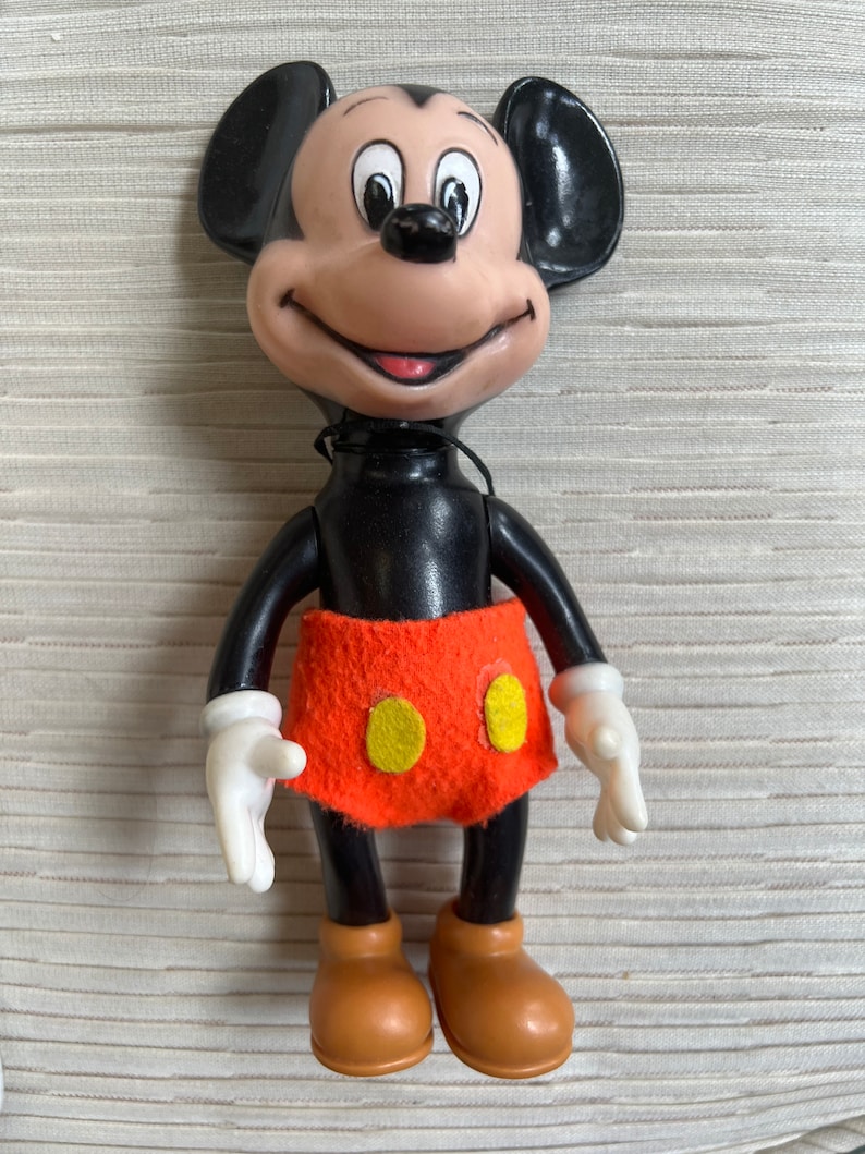 May include: A vintage Mickey Mouse figurine. The figure has a black body, large black ears, and a tan face with a smiling expression. It wears an orange skirt with two yellow circles and brown shoes. The hands are white.