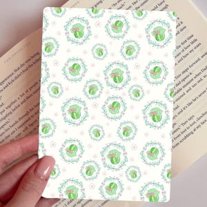 May include: A white card with a repeating pattern of green jars with pink bows and floral wreaths. The card is held in a person's hand, with a book in the background.