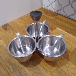 May include: A stainless steel condiment holder with three bowls and spoons. The holder has a wooden knob on top.