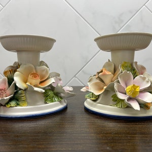 May include: Two white ceramic candle holders with floral designs. Each holder features a base with a ring of flowers, including roses, lilies, and leaves.