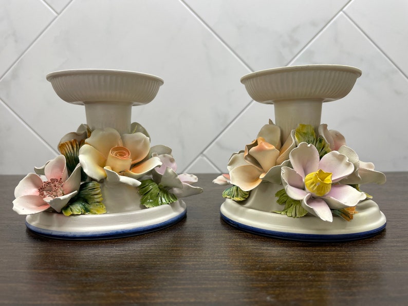 May include: Two white ceramic candle holders with floral designs. Each holder features a base with a ring of flowers, including roses, lilies, and leaves.