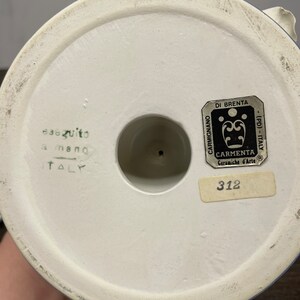 May include: White ceramic base with blue trim, featuring a label with the text "CARMIGNANO DI BRENTA - (PD) - ITALY CARMENTA Ceramiche d'Arte R 312" and the text "eseguito a mano ITALY".
