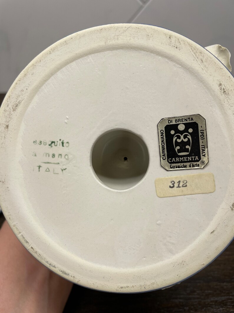 May include: White ceramic base with blue trim, featuring a label with the text "CARMIGNANO DI BRENTA - (PD) - ITALY CARMENTA Ceramiche d'Arte R 312" and the text "eseguito a mano ITALY".