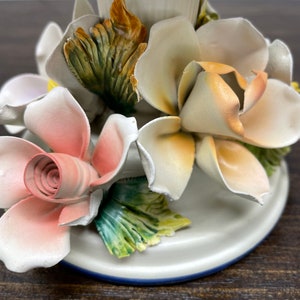 May include: A white ceramic candle holder with a floral design. The holder features three roses, one pink, one white, and one yellow, with green leaves.
