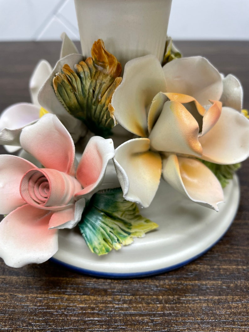 May include: A white ceramic candle holder with a floral design. The holder features three roses, one pink, one white, and one yellow, with green leaves.