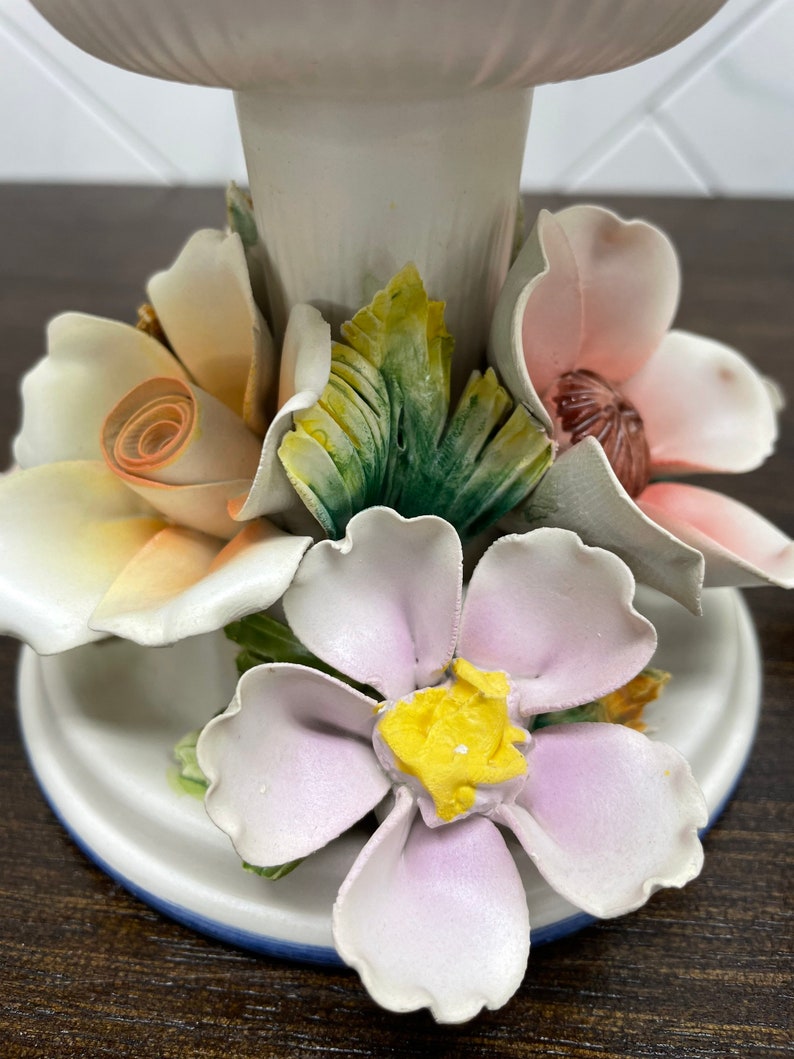 May include: A white ceramic pedestal with a floral design. The pedestal is decorated with pink, yellow, and green flowers. The flowers are three-dimensional and have a realistic appearance.