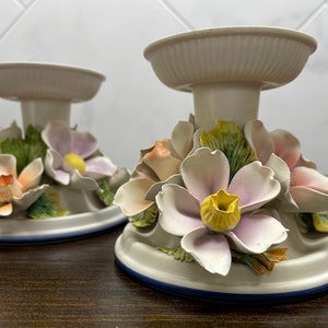 May include: Two white ceramic candle holders with floral designs. The holders feature pink, yellow, and green flowers.