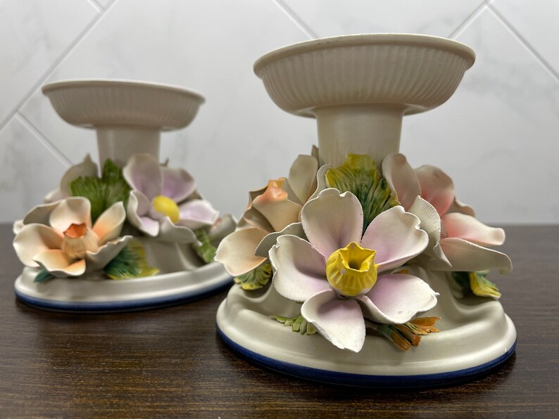 May include: Two white ceramic candle holders with floral designs. The holders feature pink, yellow, and green flowers.