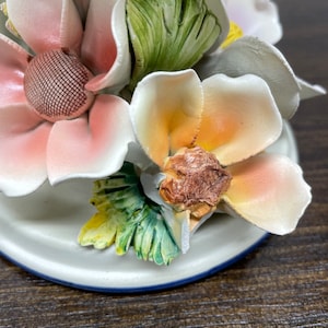 May include: A ceramic candle holder with a floral design. The holder features three pink, white, and orange flowers with green leaves. The base of the holder is white with a blue trim.
